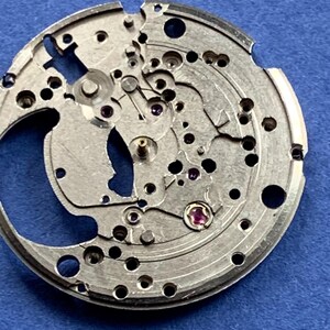 Puede incluir: Primer plano de un movimiento de reloj plateado con intrincados engranajes, agujeros y peque&ntilde;as joyas. Los componentes mec&aacute;nicos est&aacute;n dispuestos en un patr&oacute;n circular sobre un fondo azul. El mecanismo del reloj es detallado y muestra el funcionamiento interno de un reloj.
