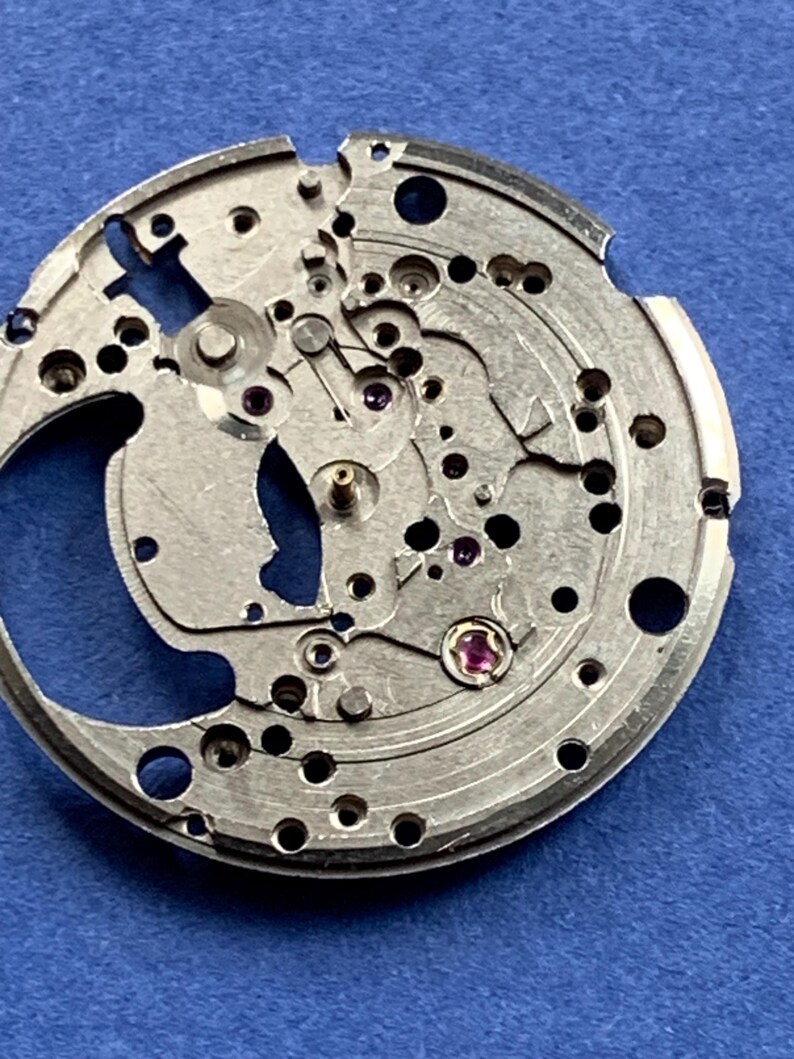Puede incluir: Primer plano de un movimiento de reloj plateado con intrincados engranajes, agujeros y peque&ntilde;as joyas. Los componentes mec&aacute;nicos est&aacute;n dispuestos en un patr&oacute;n circular sobre un fondo azul. El mecanismo del reloj es detallado y muestra el funcionamiento interno de un reloj.