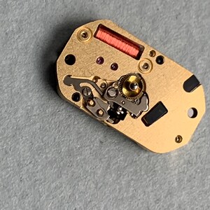 May include: Close-up of a watch movement with gold-colored metal components. The movement features intricate gears, a red coil, and small black and purple details. The watch mechanism is rectangular with rounded edges.