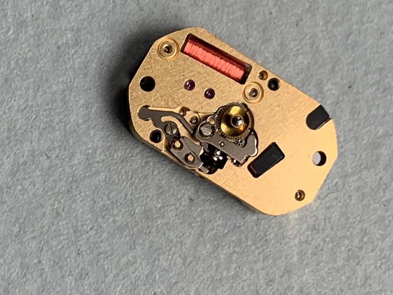 May include: Close-up of a watch movement with gold-colored metal components. The movement features intricate gears, a red coil, and small black and purple details. The watch mechanism is rectangular with rounded edges.