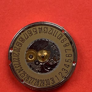 May include: Close-up of a watch movement with a gold-colored dial and silver-colored metal casing. The dial features the numbers 1 through 31, indicating the days of the month. The intricate gears and mechanisms are visible.