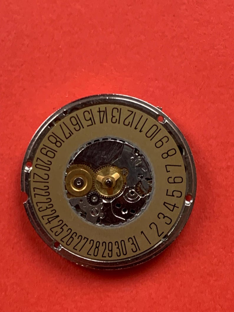 May include: Close-up of a watch movement with a gold-colored dial and silver-colored metal casing. The dial features the numbers 1 through 31, indicating the days of the month. The intricate gears and mechanisms are visible.