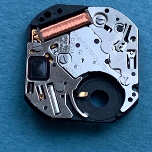 May include: Close-up of a watch movement with intricate mechanical components. The watch mechanism features copper coils, silver-colored metal parts, and the text "ORIENT WATCH CO. LTD" and "JAPAN". The watch movement is set against a blue background.