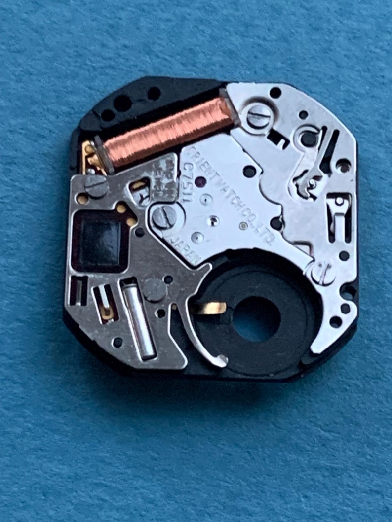 May include: Close-up of a watch movement with intricate mechanical components. The watch mechanism features copper coils, silver-colored metal parts, and the text "ORIENT WATCH CO. LTD" and "JAPAN". The watch movement is set against a blue background.