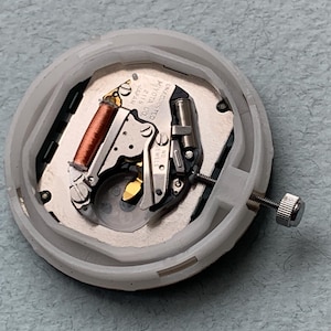 Puede incluir: Imagen detallada de un movimiento de reloj, mostrando sus intrincados componentes mec&aacute;nicos. El movimiento incluye una bobina de cobre, engranajes plateados y un tornillo. Est&aacute; alojado en una carcasa de pl&aacute;stico blanco. Se puede leer el texto "MIYOTA CO. JAPAN 2115".