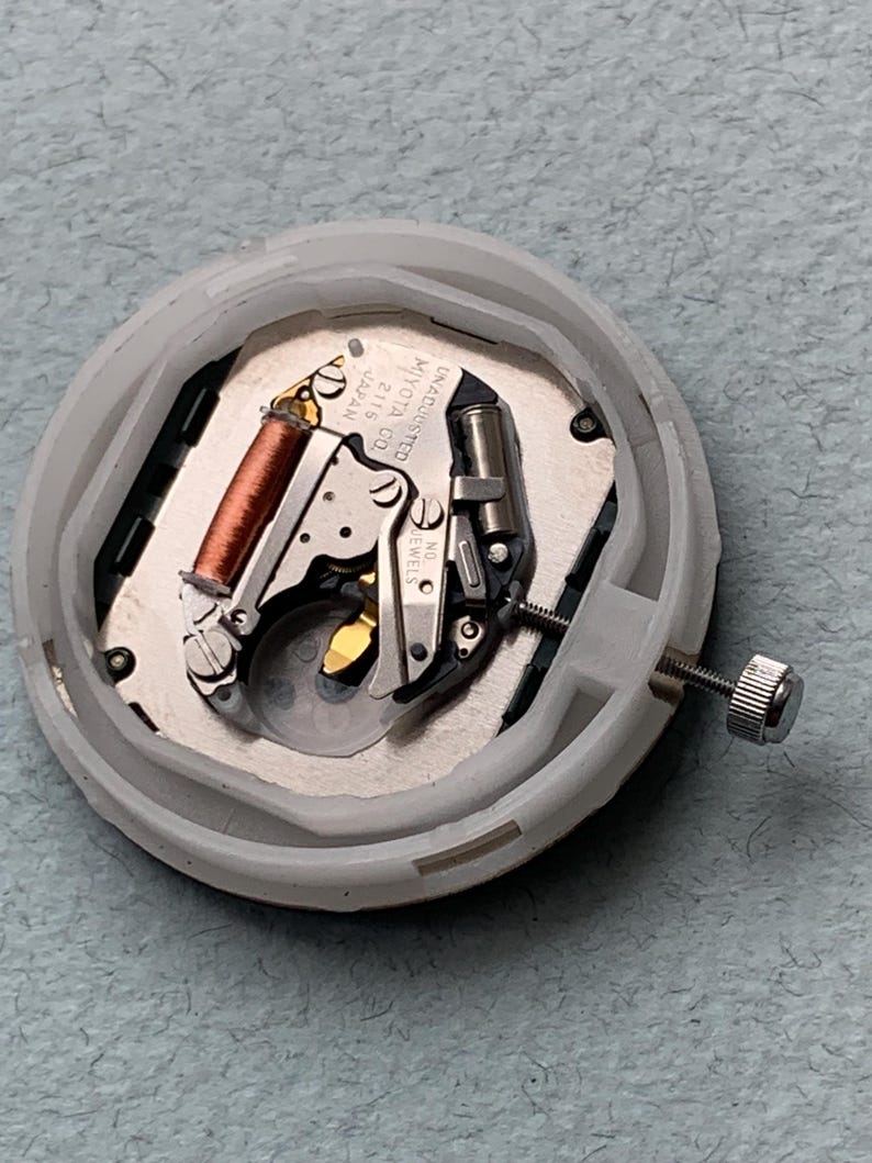 Puede incluir: Imagen detallada de un movimiento de reloj, mostrando sus intrincados componentes mec&aacute;nicos. El movimiento incluye una bobina de cobre, engranajes plateados y un tornillo. Est&aacute; alojado en una carcasa de pl&aacute;stico blanco. Se puede leer el texto "MIYOTA CO. JAPAN 2115".