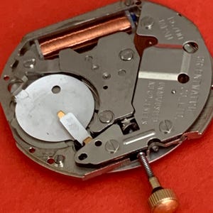 K&ouml;nnte beinhalten: Nahaufnahme eines Uhrwerks mit silbernen und kupferfarbenen Komponenten. Der Uhrenmechanismus hat eine kleine goldfarbene Krone und die Worte "ORIENT WATCH" sind sichtbar. Der Hintergrund ist einfarbig rot.