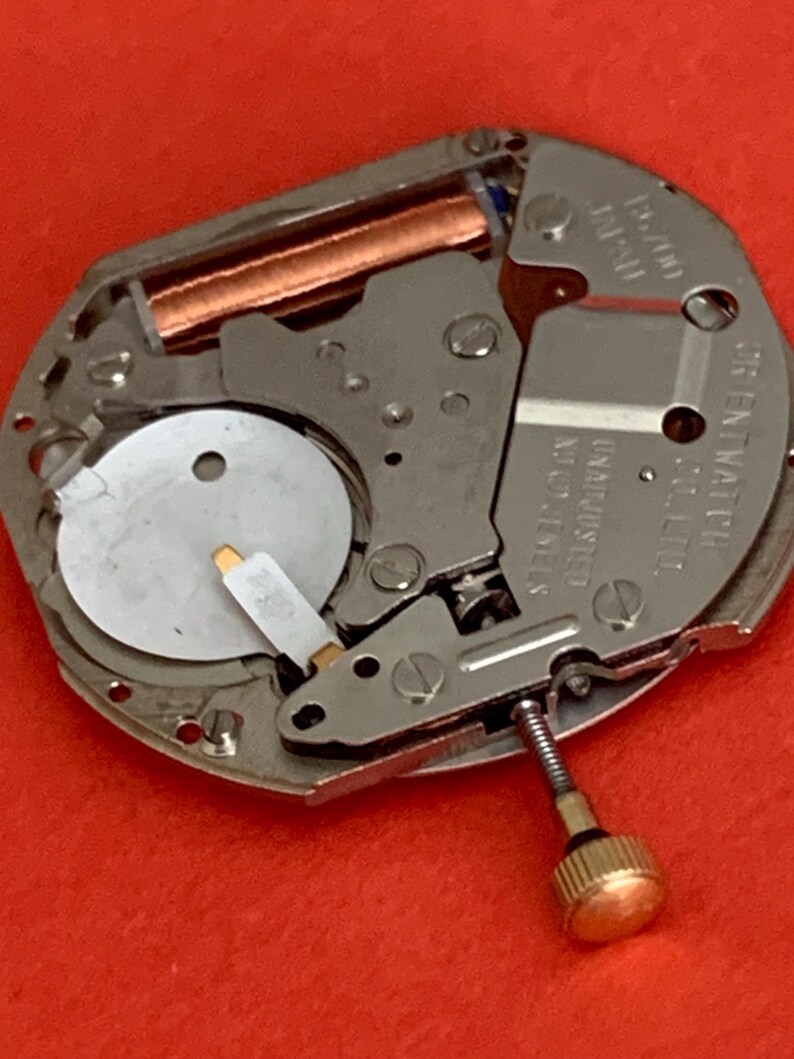 K&ouml;nnte beinhalten: Nahaufnahme eines Uhrwerks mit silbernen und kupferfarbenen Komponenten. Der Uhrenmechanismus hat eine kleine goldfarbene Krone und die Worte "ORIENT WATCH" sind sichtbar. Der Hintergrund ist einfarbig rot.
