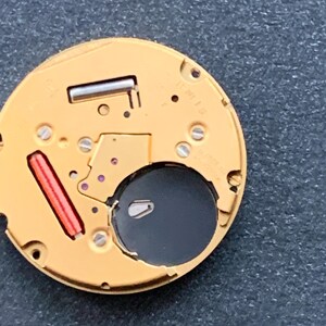 May include: Close-up of a watch movement with a gold-colored base. The movement features a black circular opening, a red rectangular component, and several small screws. The intricate design showcases the internal mechanics of a timepiece.