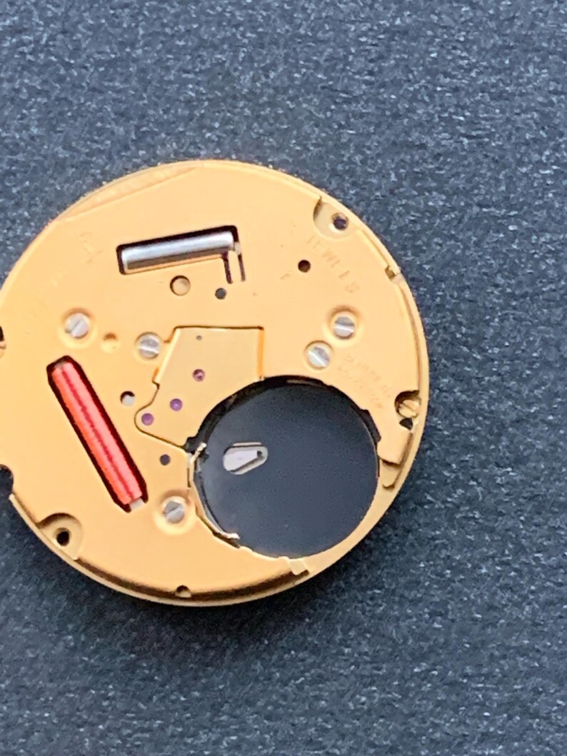 May include: Close-up of a watch movement with a gold-colored base. The movement features a black circular opening, a red rectangular component, and several small screws. The intricate design showcases the internal mechanics of a timepiece.