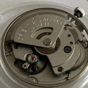 May include: Close-up of a watch movement with the text "ORIENT 21J. 46943 JAPAN" visible. The watch mechanism is silver-toned with various gears, screws, and a jewel-toned bearing. The crown is visible at the top right.
