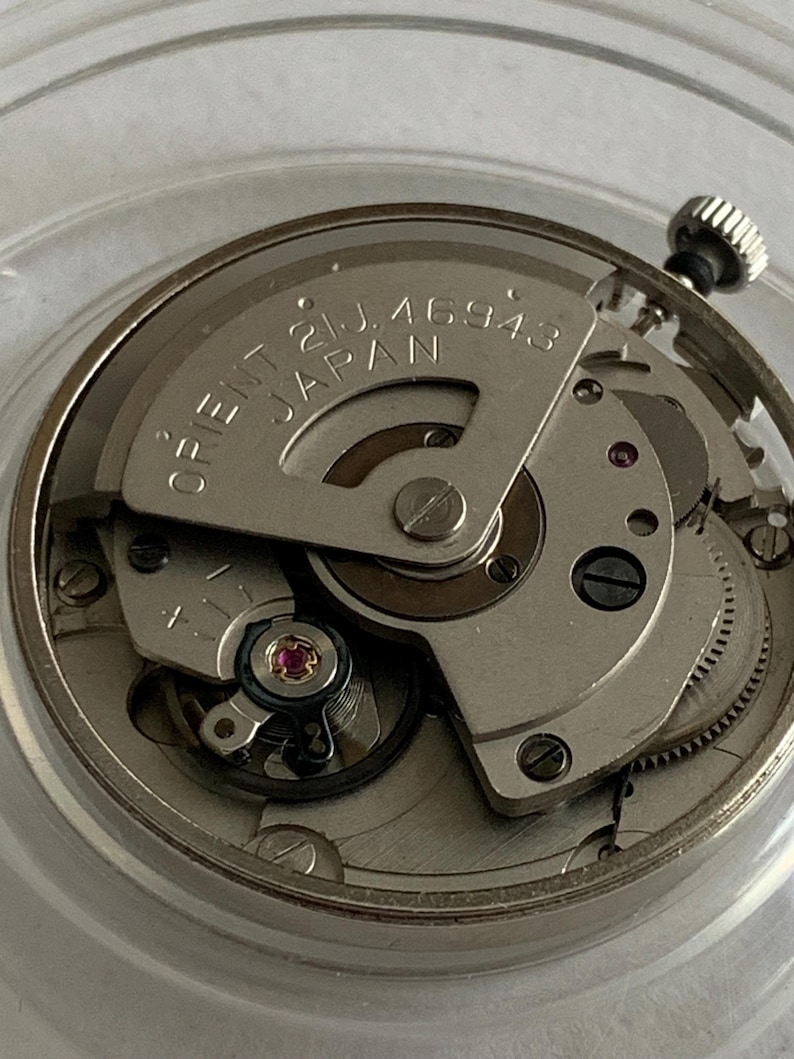 May include: Close-up of a watch movement with the text "ORIENT 21J. 46943 JAPAN" visible. The watch mechanism is silver-toned with various gears, screws, and a jewel-toned bearing. The crown is visible at the top right.