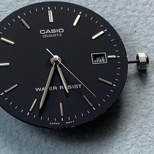 Puede incluir: Una esfera de reloj Casio Quartz negra con manecillas e indicadores plateados. La esfera tiene una ventana de fecha a las 3 en punto y una corona plateada. Las palabras "CASIO QUARTZ" y "WATER RESIST" est&aacute;n impresas en la esfera.