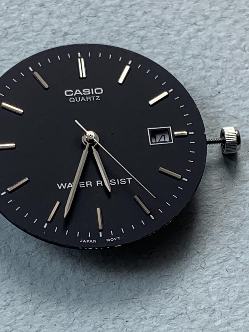 Puede incluir: Una esfera de reloj Casio Quartz negra con manecillas e indicadores plateados. La esfera tiene una ventana de fecha a las 3 en punto y una corona plateada. Las palabras "CASIO QUARTZ" y "WATER RESIST" est&aacute;n impresas en la esfera.
