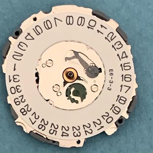 May include: A watch movement component with a white dial featuring black numerals from 1 to 31, indicating the date. The component has a complex mechanical design with gears and a small green piece. The text "E6-3-3" is visible.