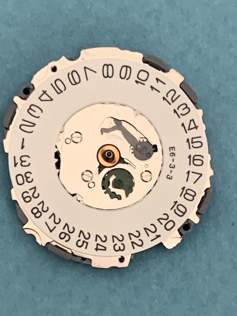 May include: A watch movement component with a white dial featuring black numerals from 1 to 31, indicating the date. The component has a complex mechanical design with gears and a small green piece. The text "E6-3-3" is visible.