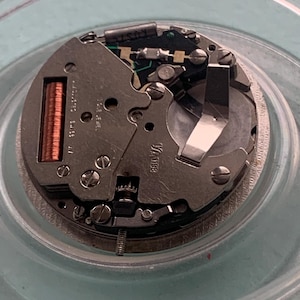 Op de afbeelding: Close-up van een uurwerk met ingewikkelde mechanische componenten. De zilverkleurige metalen onderdelen omvatten tandwielen, schroeven en een spoel. Het uurwerk is geplaatst in een heldere, cirkelvormige container.