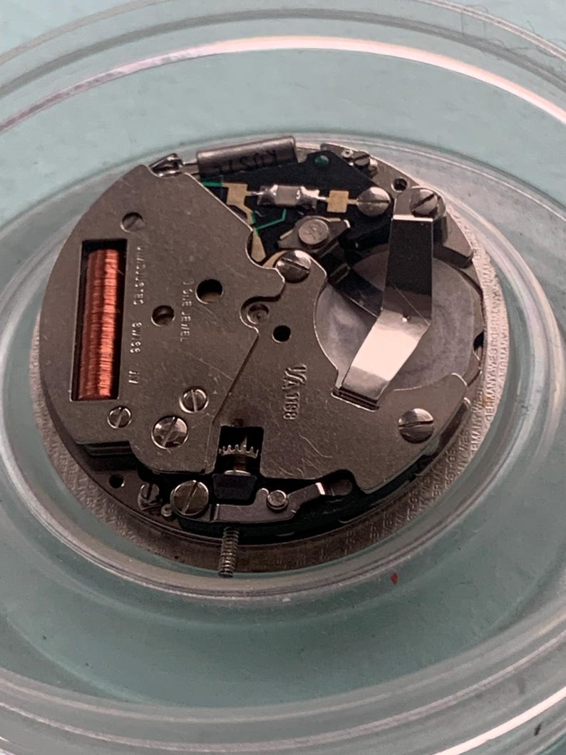 Op de afbeelding: Close-up van een uurwerk met ingewikkelde mechanische componenten. De zilverkleurige metalen onderdelen omvatten tandwielen, schroeven en een spoel. Het uurwerk is geplaatst in een heldere, cirkelvormige container.
