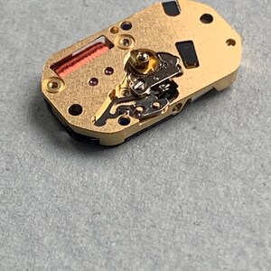 May include: A close-up of a watch movement, featuring a gold-colored base with intricate gears, a red coil, and small jewels. The rectangular component has several holes and black accents, showcasing the detailed mechanics of a timepiece.