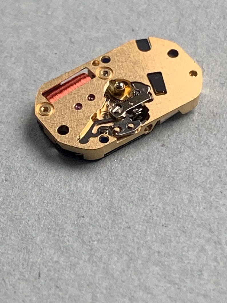 May include: A close-up of a watch movement, featuring a gold-colored base with intricate gears, a red coil, and small jewels. The rectangular component has several holes and black accents, showcasing the detailed mechanics of a timepiece.
