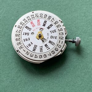 May include: A watch movement with a white dial showing the days of the week and the date. The dial has black numerals and text, with the day of the week in English and German. The date numbers are around the outer edge of the dial.