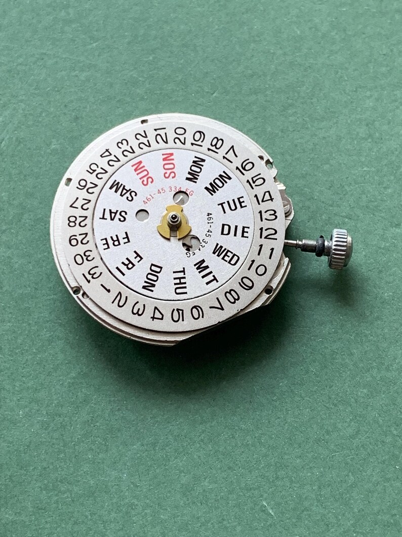 May include: A watch movement with a white dial showing the days of the week and the date. The dial has black numerals and text, with the day of the week in English and German. The date numbers are around the outer edge of the dial.