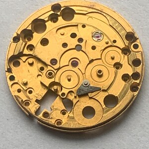 May include: Close-up of a gold-colored watch movement. The intricate mechanism features numerous gears, holes, and a small, jewel-like component. The circular design showcases the detailed craftsmanship of a vintage timepiece.