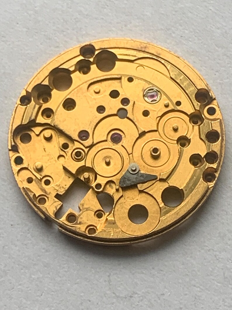 May include: Close-up of a gold-colored watch movement. The intricate mechanism features numerous gears, holes, and a small, jewel-like component. The circular design showcases the detailed craftsmanship of a vintage timepiece.