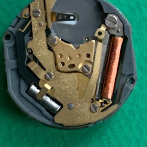 May include: Close-up of a watch movement with gold-colored gears, a copper coil, and a silver battery. The movement is set in a gray plastic casing. The image is taken against a green background.
