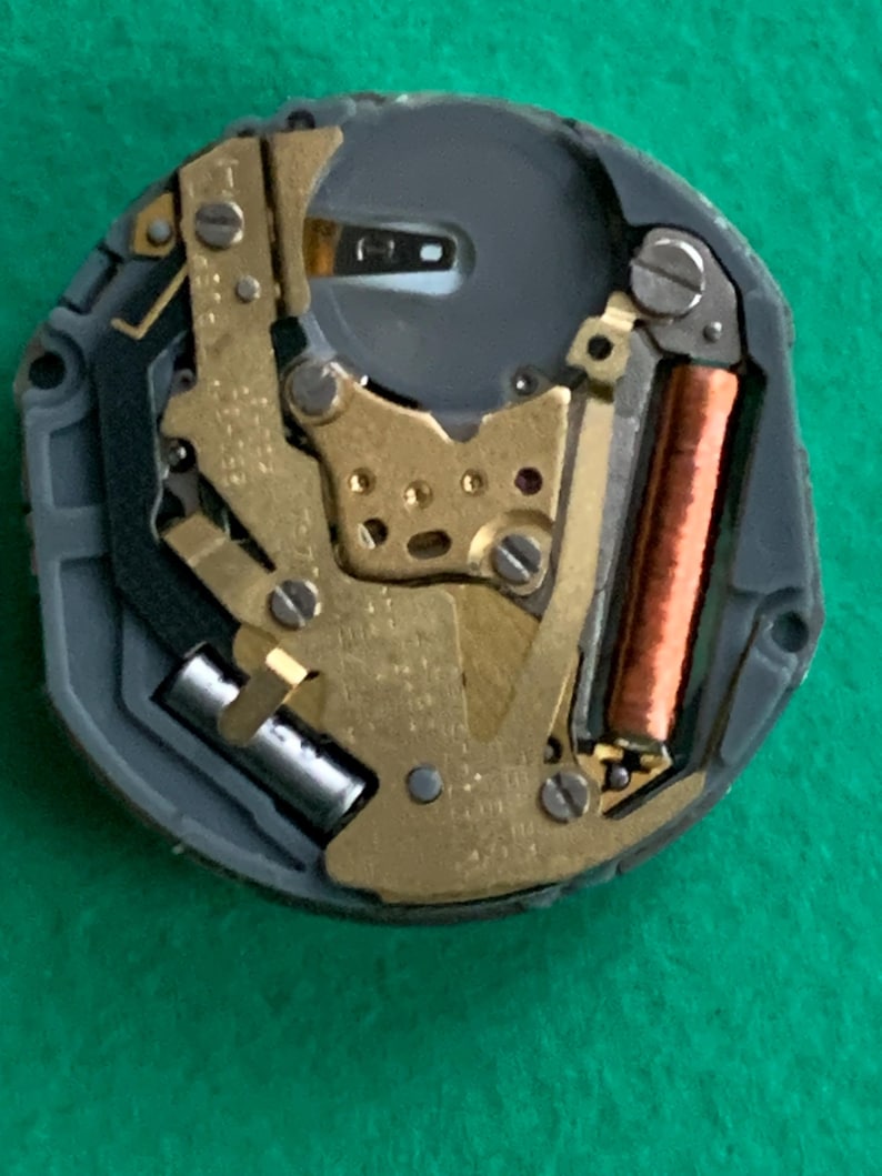 May include: Close-up of a watch movement with gold-colored gears, a copper coil, and a silver battery. The movement is set in a gray plastic casing. The image is taken against a green background.
