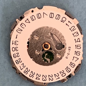 May include: Close-up of a watch movement with a white date dial and black numbers. The dial has a central mechanism with a green jewel and is surrounded by a metal frame. The text "E6-3-3" is visible.