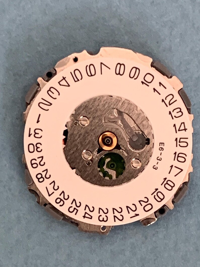 May include: Close-up of a watch movement with a white date dial and black numbers. The dial has a central mechanism with a green jewel and is surrounded by a metal frame. The text "E6-3-3" is visible.