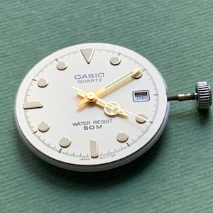 K&ouml;nnte beinhalten: Ein Casio Quartz Zifferblatt mit cremefarbenem Zifferblatt, goldenen Zeigern und goldenen Stundenmarkierungen. Das Zifferblatt zeigt "CASIO QUARTZ" und "WATER RESIST 50 M". Das Datum wird bei 3 Uhr angezeigt. Klassisches Design.
