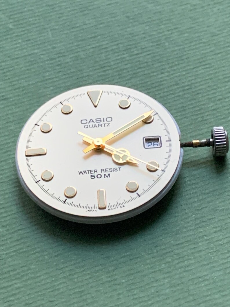 K&ouml;nnte beinhalten: Ein Casio Quartz Zifferblatt mit cremefarbenem Zifferblatt, goldenen Zeigern und goldenen Stundenmarkierungen. Das Zifferblatt zeigt "CASIO QUARTZ" und "WATER RESIST 50 M". Das Datum wird bei 3 Uhr angezeigt. Klassisches Design.
