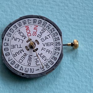 K&ouml;nnte beinhalten: Ein Uhrenkalenderzifferblatt mit wei&szlig;em Zifferblatt, das Wochentage in mehreren Sprachen und Zahlen anzeigt. Das Zifferblatt ist von einem grauen Ring umgeben und hat eine goldfarbene Einstellschraube.