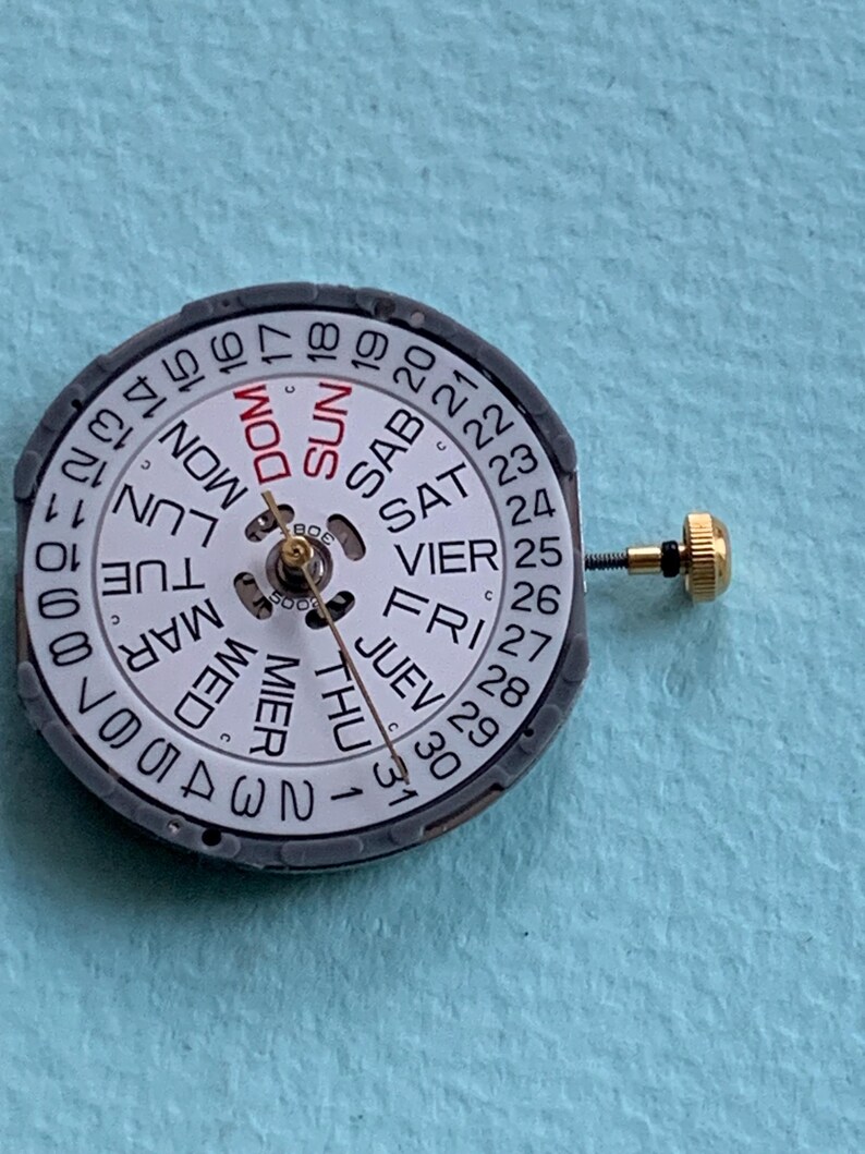 K&ouml;nnte beinhalten: Ein Uhrenkalenderzifferblatt mit wei&szlig;em Zifferblatt, das Wochentage in mehreren Sprachen und Zahlen anzeigt. Das Zifferblatt ist von einem grauen Ring umgeben und hat eine goldfarbene Einstellschraube.