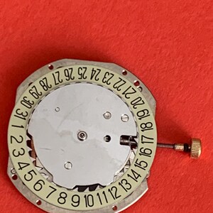 K&ouml;nnte beinhalten: Ein Uhrwerk mit silberfarbenem Zifferblatt und kreisf&ouml;rmiger Datumsanzeige. Die Datumszahlen sind in Schwarz auf einem hellgr&uuml;nen Ring gedruckt. Eine kleine goldfarbene Krone ist seitlich angebracht. Der Hintergrund ist einfarbig rot.