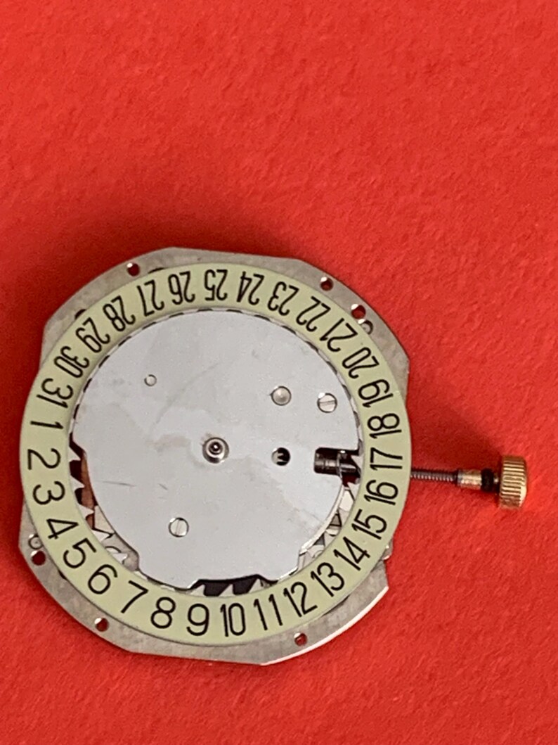 K&ouml;nnte beinhalten: Ein Uhrwerk mit silberfarbenem Zifferblatt und kreisf&ouml;rmiger Datumsanzeige. Die Datumszahlen sind in Schwarz auf einem hellgr&uuml;nen Ring gedruckt. Eine kleine goldfarbene Krone ist seitlich angebracht. Der Hintergrund ist einfarbig rot.
