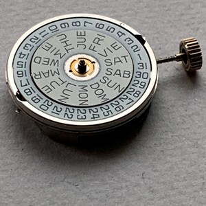 Puede incluir: Un movimiento de reloj con una esfera gris claro que muestra los d&iacute;as de la semana y la fecha. La esfera tiene un anillo exterior plateado con los n&uacute;meros 1-31. Un peque&ntilde;o componente central dorado y una corona dorada son visibles.