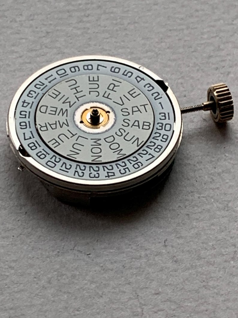 Puede incluir: Un movimiento de reloj con una esfera gris claro que muestra los d&iacute;as de la semana y la fecha. La esfera tiene un anillo exterior plateado con los n&uacute;meros 1-31. Un peque&ntilde;o componente central dorado y una corona dorada son visibles.