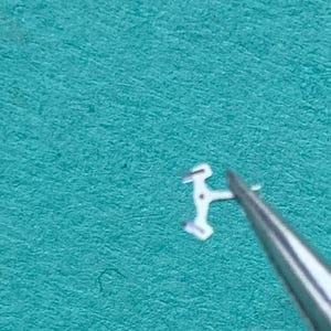 May include: A tiny, silver-colored watch component is held by tweezers against a teal background. The component has a unique shape with multiple angles and small protrusions, suggesting it is a delicate part of a watch mechanism.