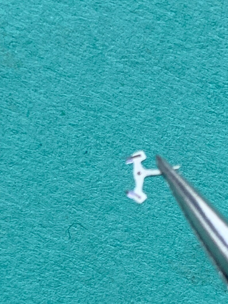 May include: A tiny, silver-colored watch component is held by tweezers against a teal background. The component has a unique shape with multiple angles and small protrusions, suggesting it is a delicate part of a watch mechanism.