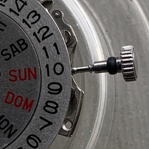 Pu&ograve; includere: Primo piano di un movimento di orologio con quadrante e lancette argentate. Il quadrante mostra i giorni della settimana in rosso e nero, con numeri attorno al bordo. Una corona argentata &egrave; visibile a destra.