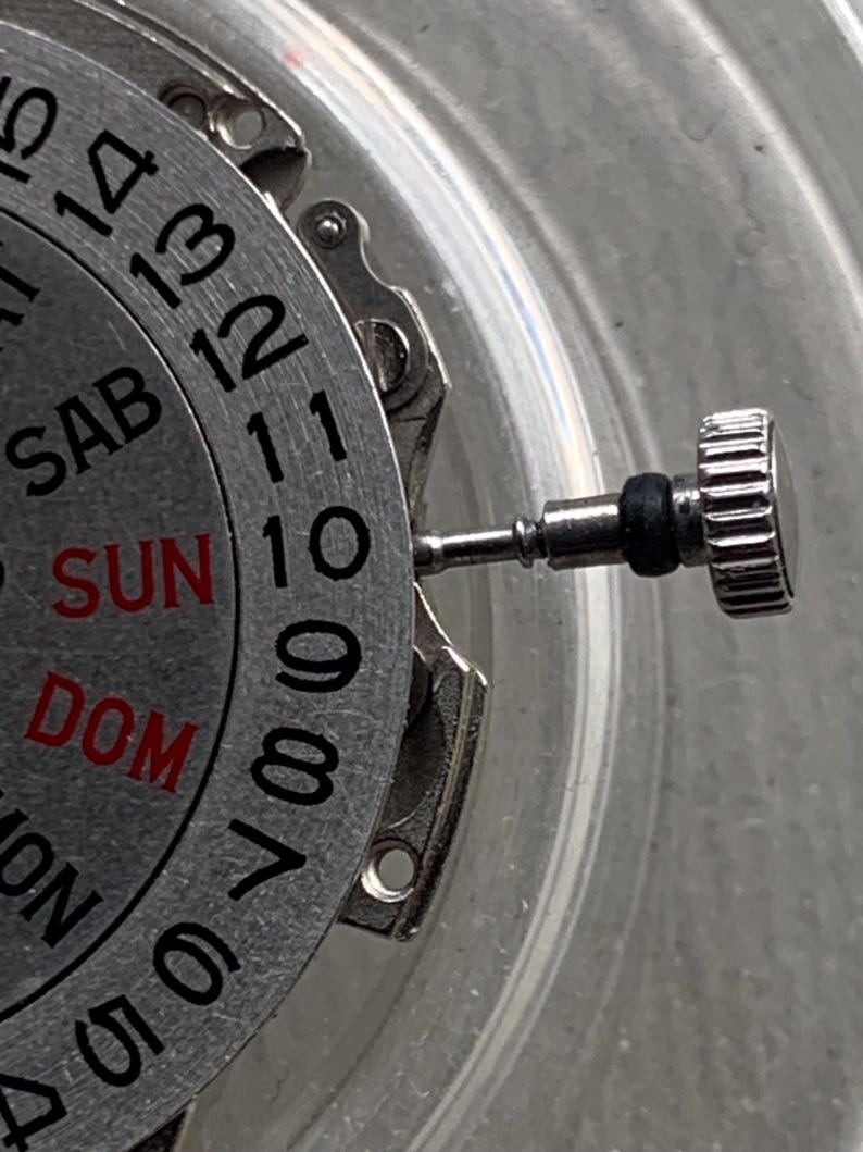 Pu&ograve; includere: Primo piano di un movimento di orologio con quadrante e lancette argentate. Il quadrante mostra i giorni della settimana in rosso e nero, con numeri attorno al bordo. Una corona argentata &egrave; visibile a destra.