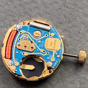 May include: Close-up of a watch movement with a blue circuit board and gold-colored components. The watch movement has the text "ZENITH" and "SWISS" printed on it. The movement also has a red coil and a screw.