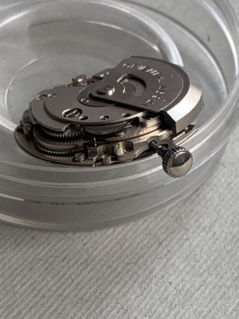 Pu&ograve; includere: Primo piano di un movimento di orologio, con ingranaggi e componenti intricati. Le parti metalliche sono argentate, con la scritta "ORIENT" visibile su una placca circolare. Il movimento &egrave; all'interno di un contenitore trasparente e rotondo.