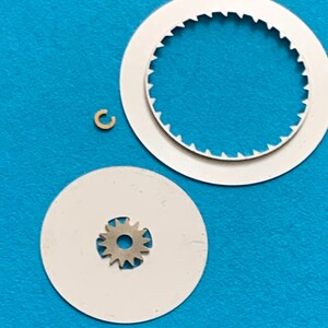 May include: A collection of watch parts, including two white circular pieces, one with a toothed inner edge, and a small, gold-colored, C-shaped component. A small silver gear is centered on a white disc. The parts are set against a bright blue background.