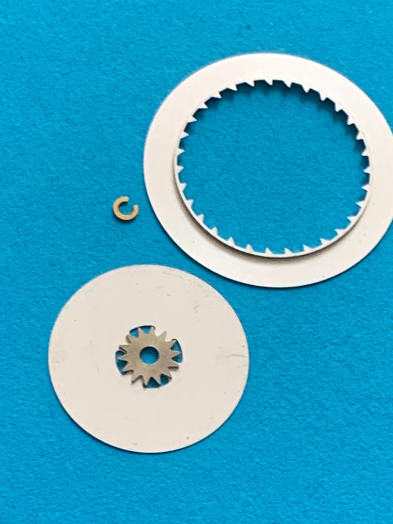 May include: A collection of watch parts, including two white circular pieces, one with a toothed inner edge, and a small, gold-colored, C-shaped component. A small silver gear is centered on a white disc. The parts are set against a bright blue background.