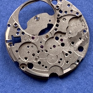 Puede incluir: Primer plano de un movimiento de reloj plateado con intrincados engranajes, agujeros y peque&ntilde;as joyas de rub&iacute;. El mecanismo circular tiene una gran abertura y varios componentes con forma. El fondo es azul liso.