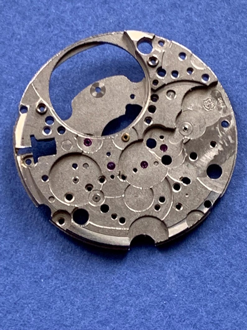 Puede incluir: Primer plano de un movimiento de reloj plateado con intrincados engranajes, agujeros y peque&ntilde;as joyas de rub&iacute;. El mecanismo circular tiene una gran abertura y varios componentes con forma. El fondo es azul liso.